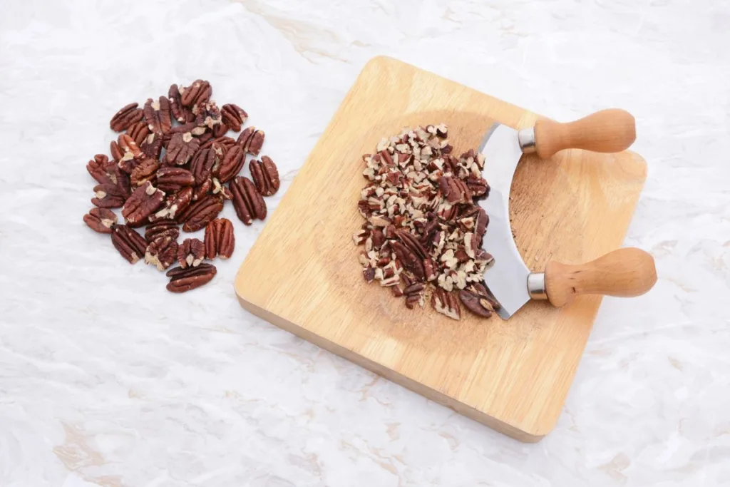 Whole toasted pecans with a rocking knife chopping the nuts on a board.