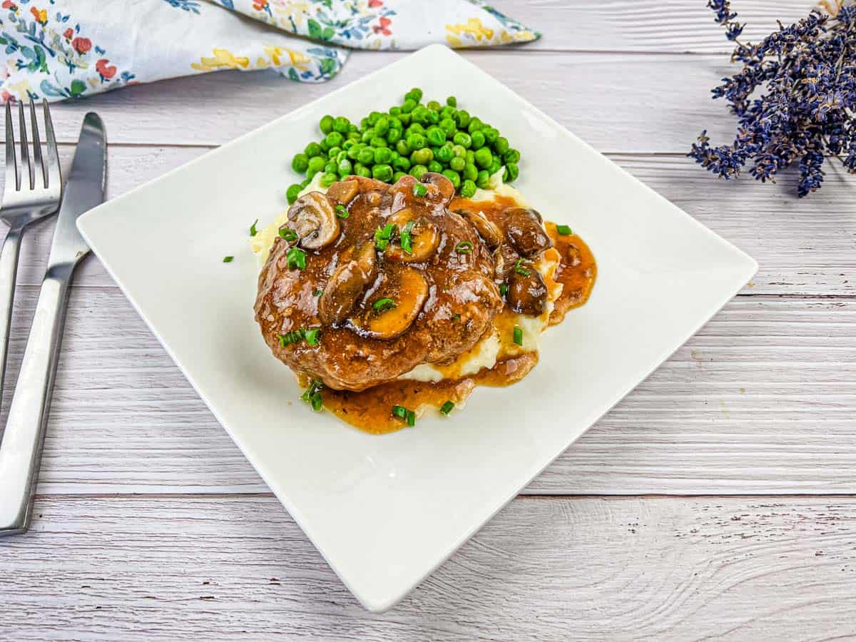 A plate of Salisbury steak with gravy on mashed potatoes, and a side of peas on a white square dish.