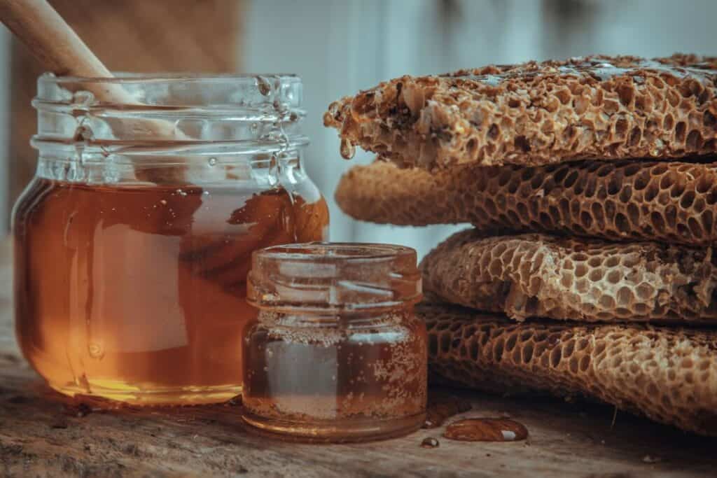 Close-Up Of Honey Dripping From Fresh Honeycombs On Glass Jar With Wooden Honey Dipper Stick On Old Wooden Table.