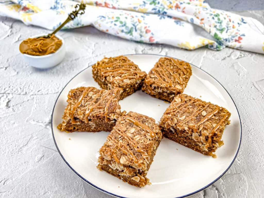 5 Biscoff Cookie Butter Streusel Bars on a white plate with a bowl of cookie butter in the background.