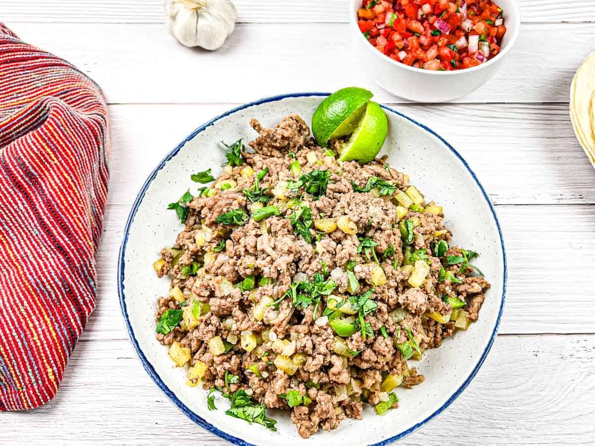 A plate of cooked ground beef mixed with diced vegetables and garnished with lime wedges and chopped herbs. A red striped cloth is placed beside the plate.