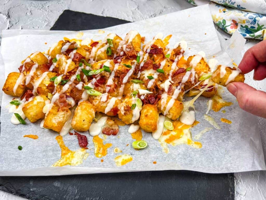 A hand-serving cheesy tater tots topped with bacon bits and green onions on white parchment paper.