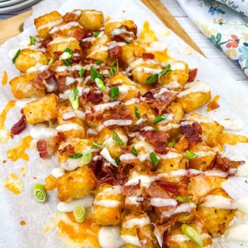 A serving of loaded tater tots topped with melted cheese, bacon bits, and green onions, and drizzled with Alfredo sauce on a wooden board.