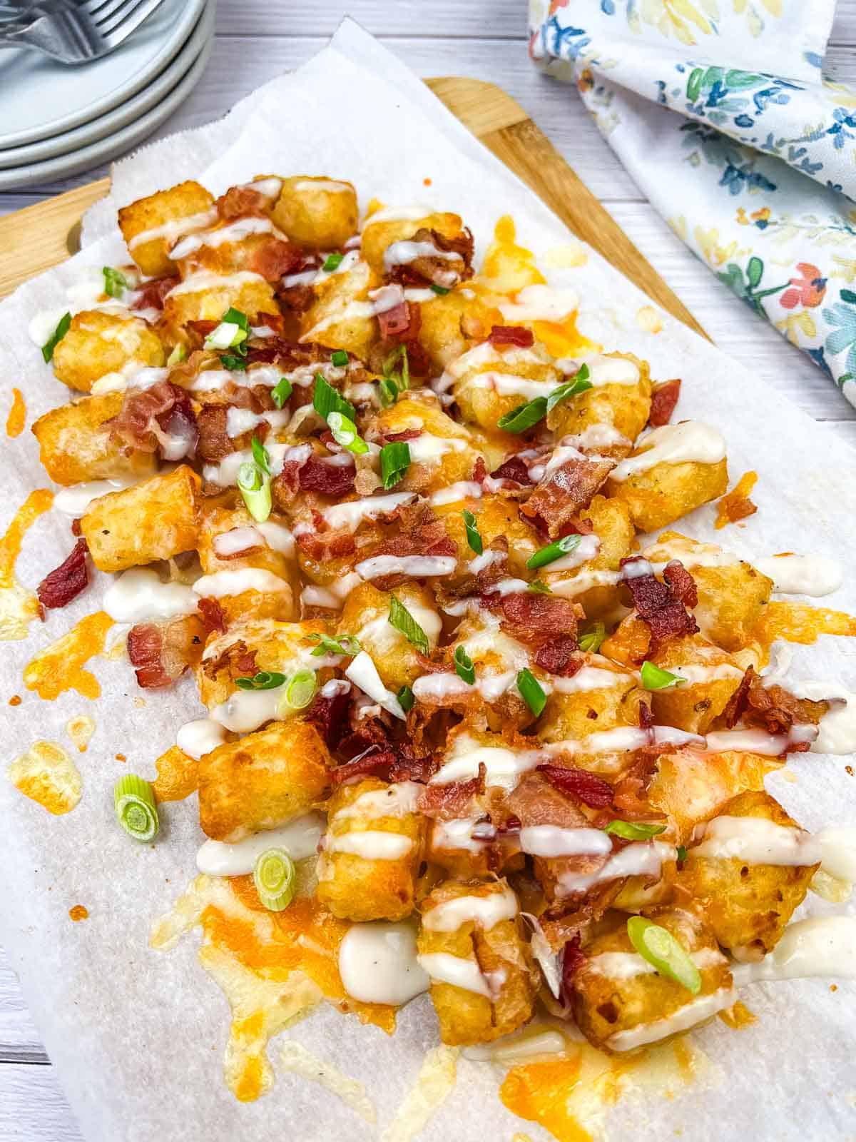 A serving of loaded tater tots topped with melted cheese, bacon bits, and green onions, and drizzled with Alfredo sauce on a wooden board.
