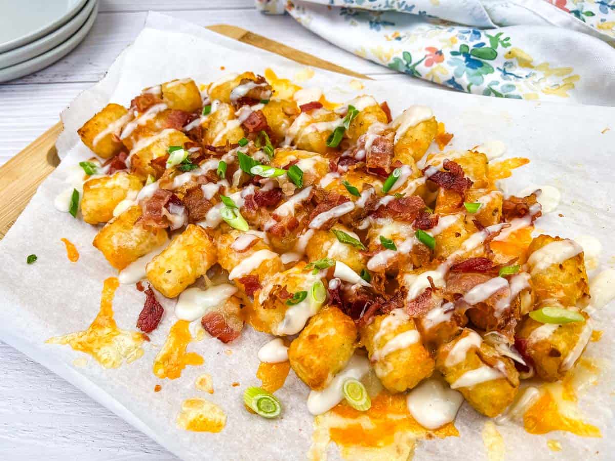 A serving of loaded tater tots topped with melted cheese, bacon bits, and green onions, and drizzled with Alfredo sauce on a wooden board.