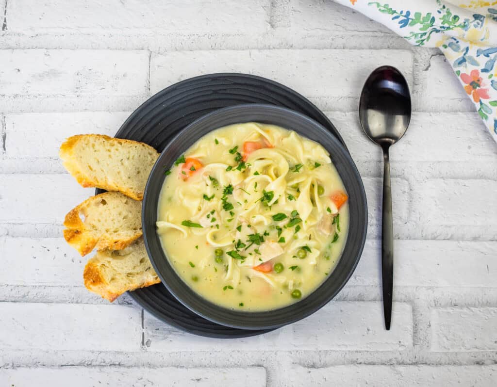 Top-down shot of a Creamy Chicken Noodle Soup in a black bowl served together with slices of bread on a plate and spoon.