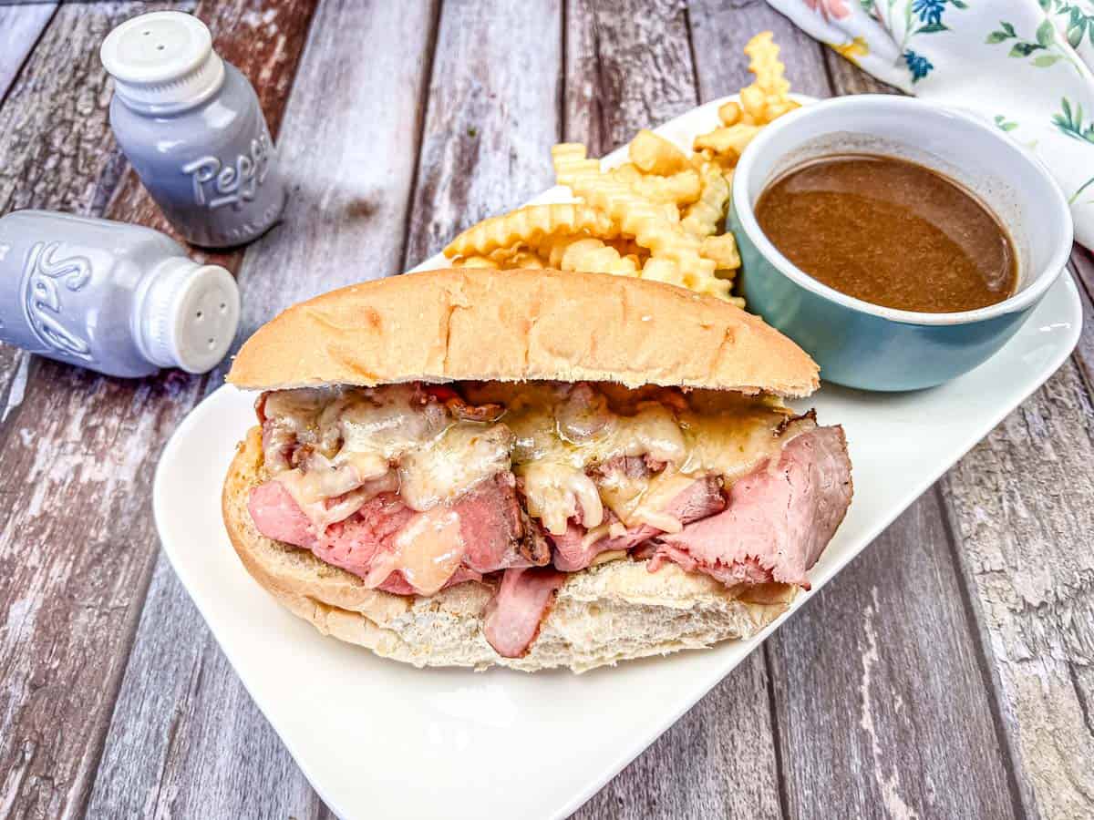 A roast beef sandwich with melted cheese on a hoagie roll, accompanied by crinkle-cut fries and a bowl of brown dipping sauce on the side.