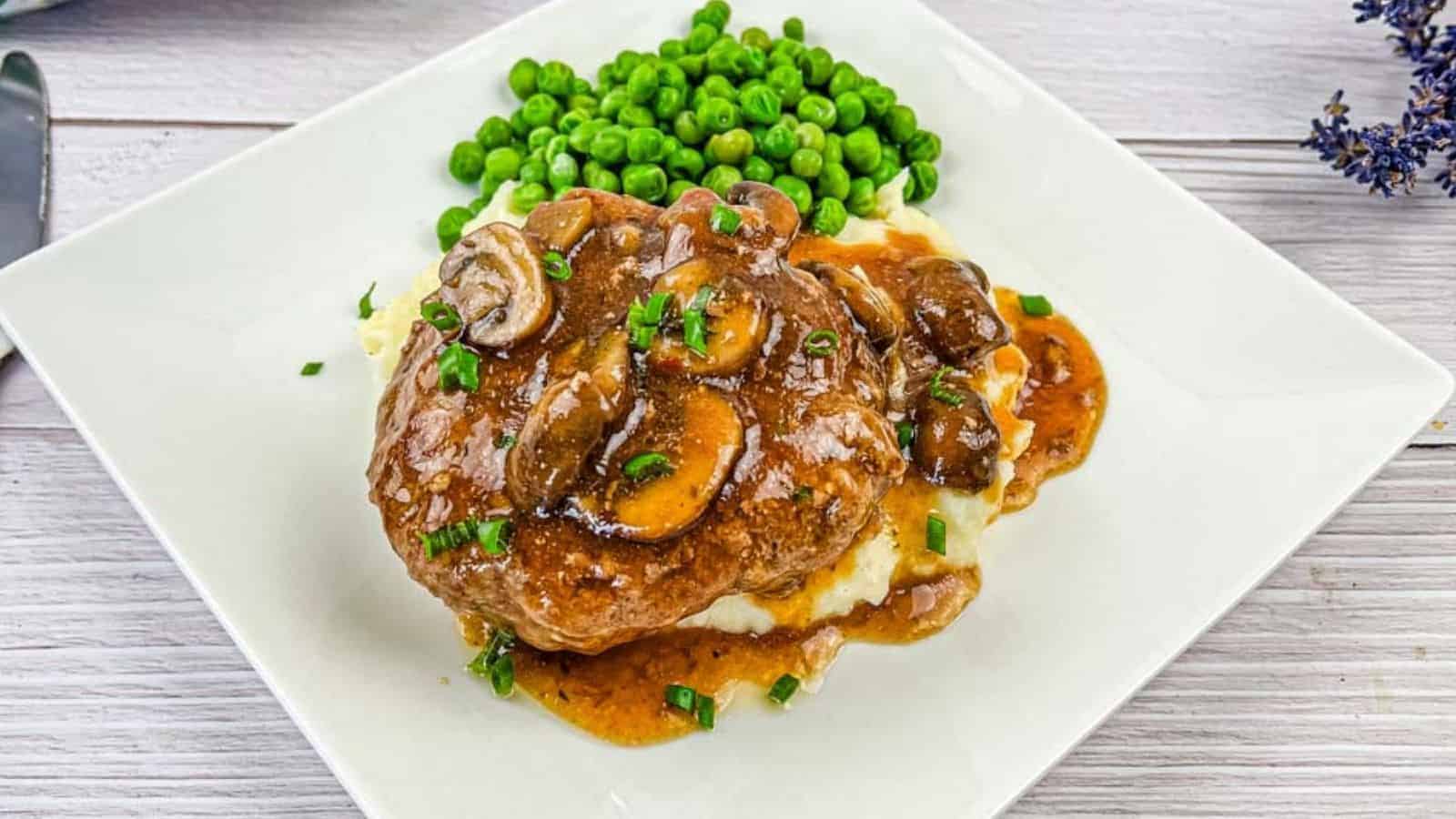 A white plate with Salisbury steak topped with mushroom gravy on mashed potatoes, accompanied by a side of green peas.