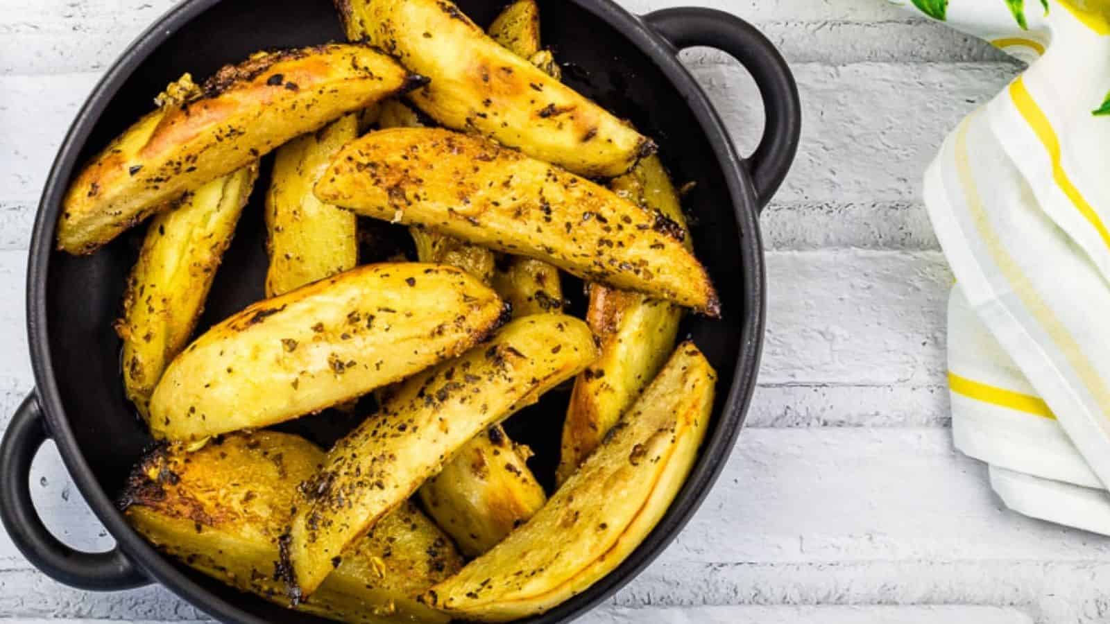 Round black dish filled with seasoned roasted potato wedges, placed on a white wooden surface alongside a white and yellow striped cloth napkin.