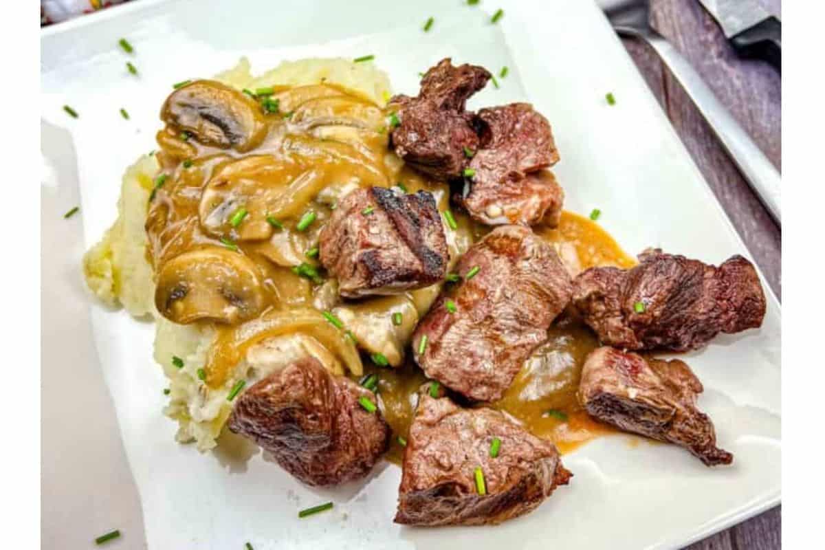 A plate of Copycat Roadhouse beef tips with gravy over mashed potatoes, garnished with sliced mushrooms and chopped chives.