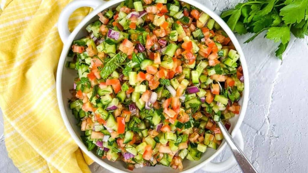 A bowl of chopped vegetable salad with cucumbers, tomatoes, red onions, and parsley, next to a yellow checkered cloth and fresh parsley.