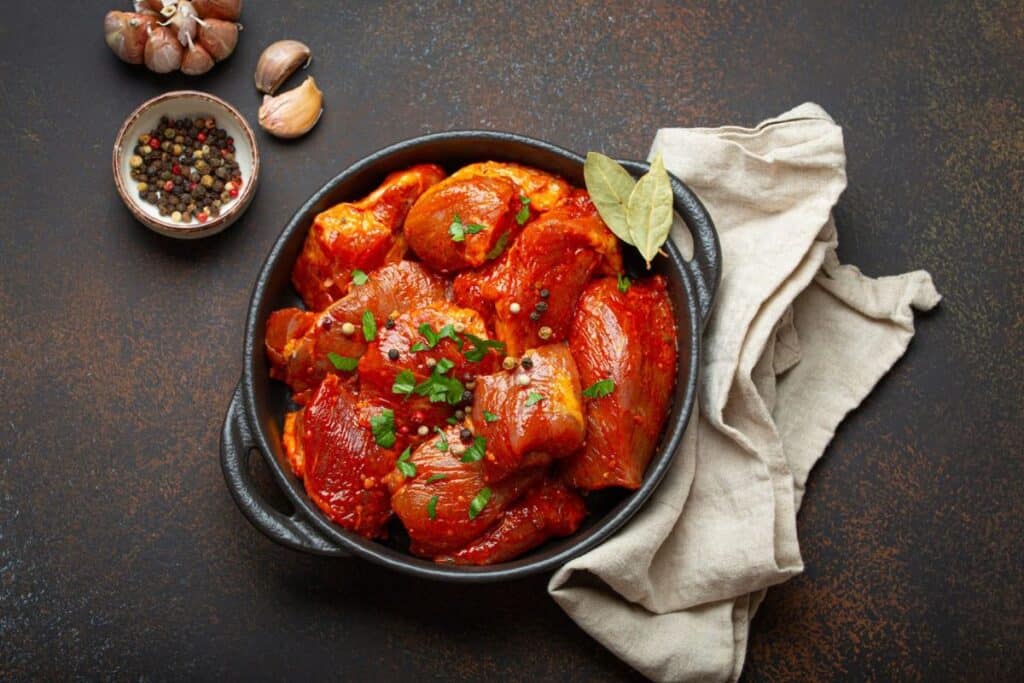 Raw Uncooked Chopped Pieces Of Meat Marinated With Seasonings And Parsley In Black Casserole Dish.