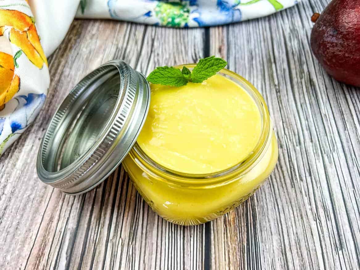 A jar of mango curd next to a mango and a mint leaf.