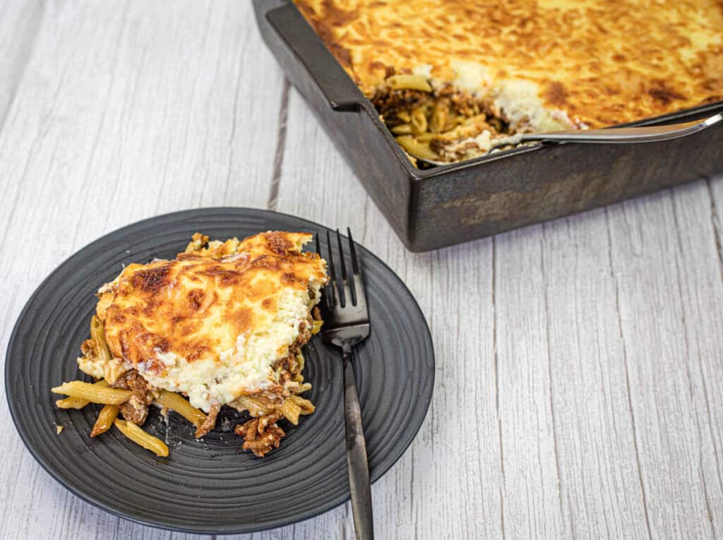 A slice of Pastitsio on a black plate beside the tray of Pastitsio.