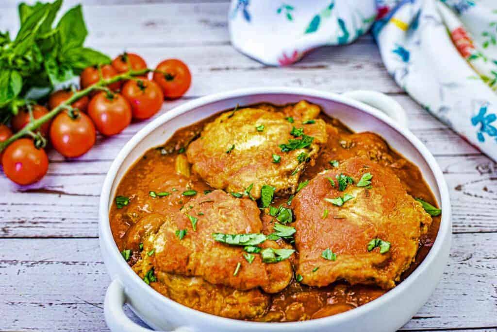 A white dish filled with pieces of Chicken Cacciatore in a red sauce, garnished with chopped herbs.