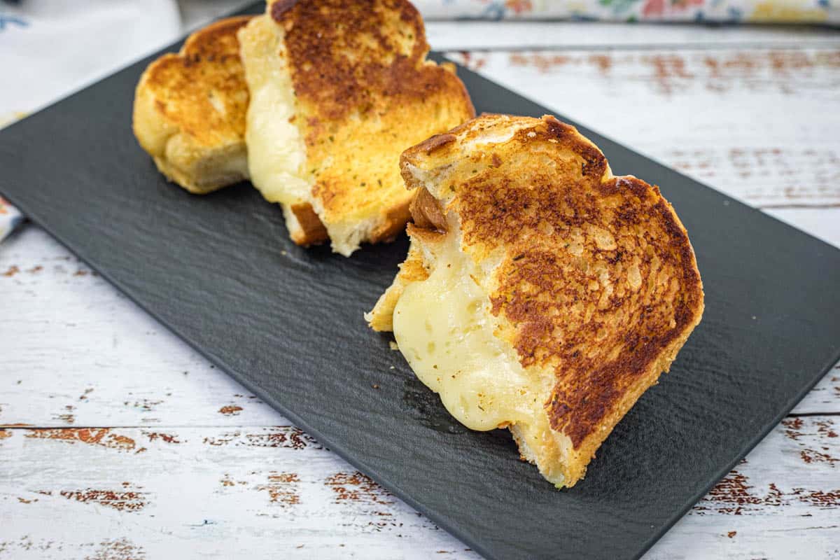 Three garlic bread grilled cheese on a black slate plate on a white wooden surface.