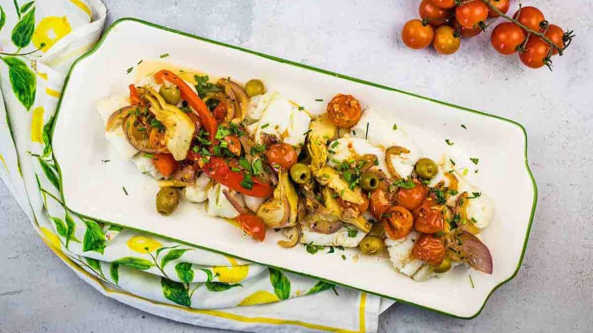 A rectangular plate with Mediterranean Cod placed on a lemon-patterned cloth. A vine of cherry tomatoes is beside the plate.