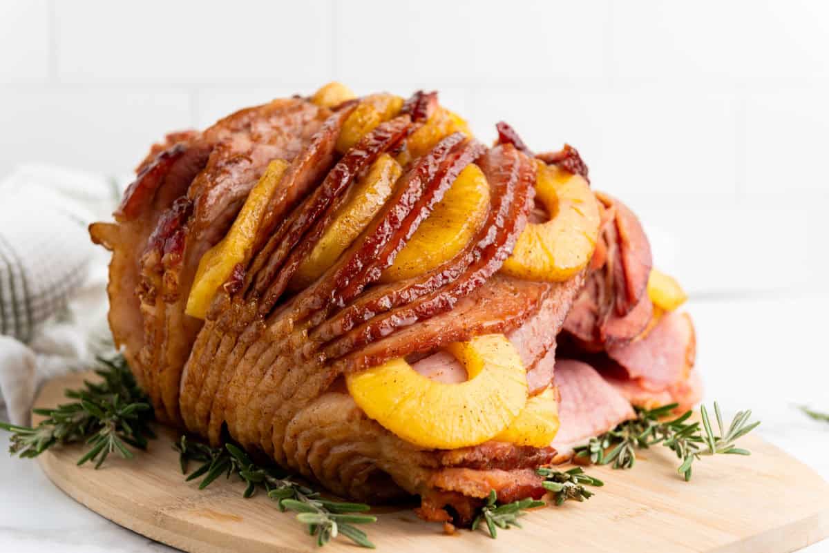 A Brown Sugar Pineapple Ham is placed on a wooden board, garnished with sprigs of rosemary.