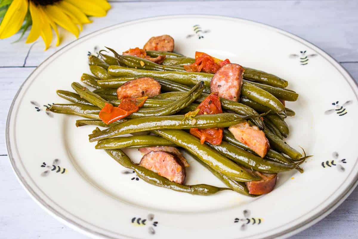 A plate of cooked Southern Green Beans with sliced sausages and tomato pieces on a white dish, placed near a sunflower.