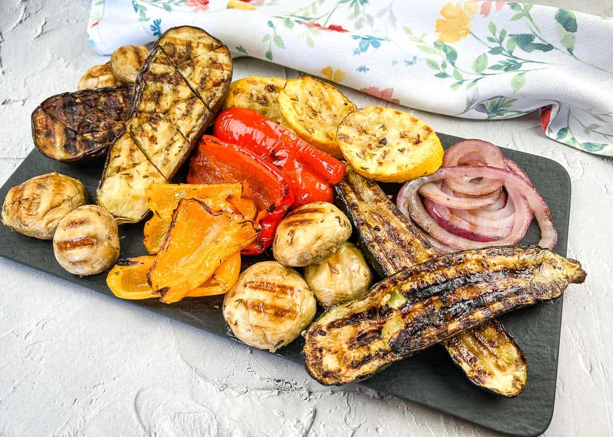 Grilled vegetables including zucchini, mushrooms, red onions, peppers, and squash, arranged on a rectangular black platter.