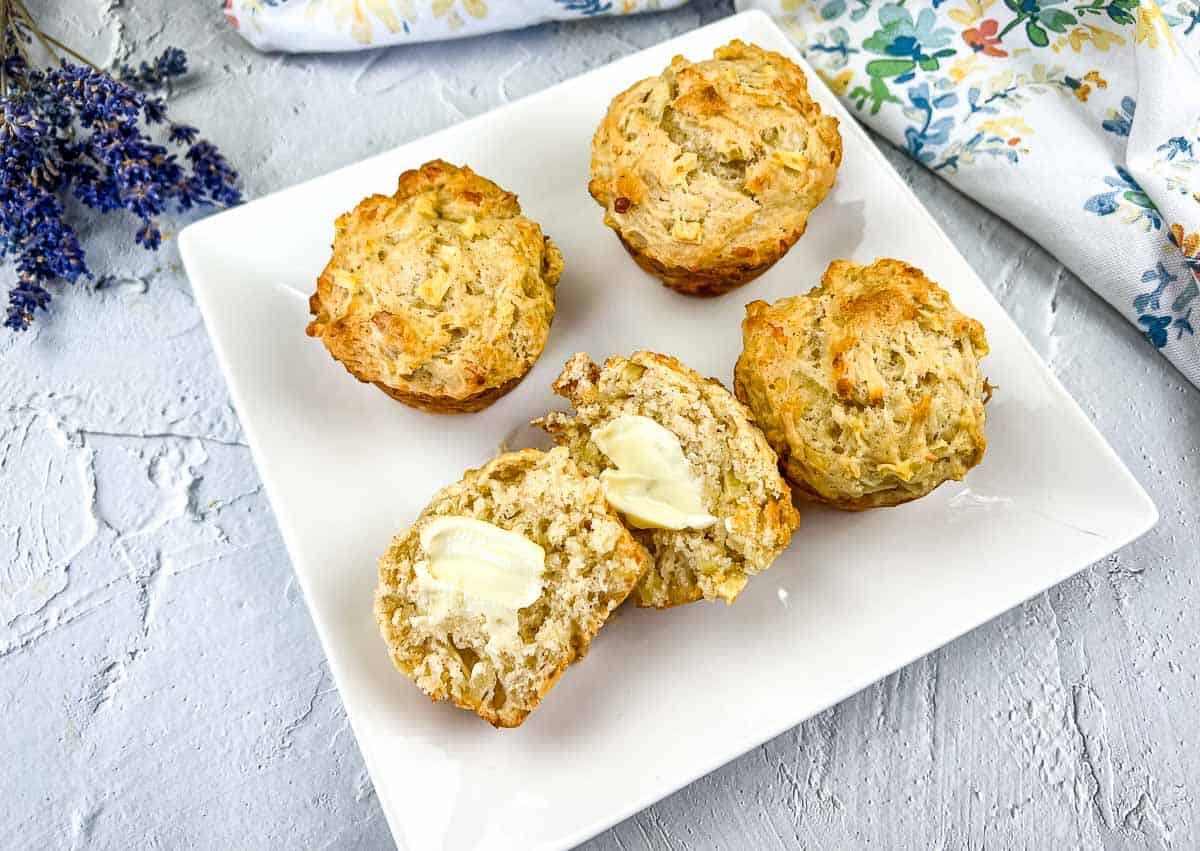 Apple cheddar muffins on a white plate, with one muffin cut open and spread with butter, showing the moist, cheesy interior.