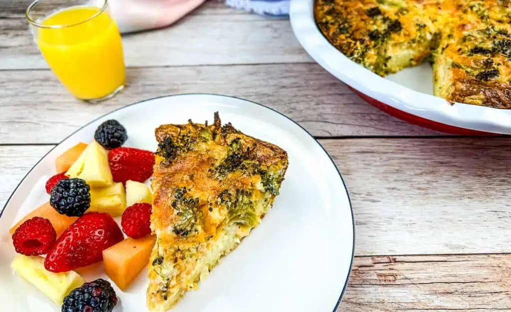 A slice of Broccoli 3-Cheese Impossible Quiche on a white plate with a side of mixed fruit. A full quiche and glass of orange juice are in the background.