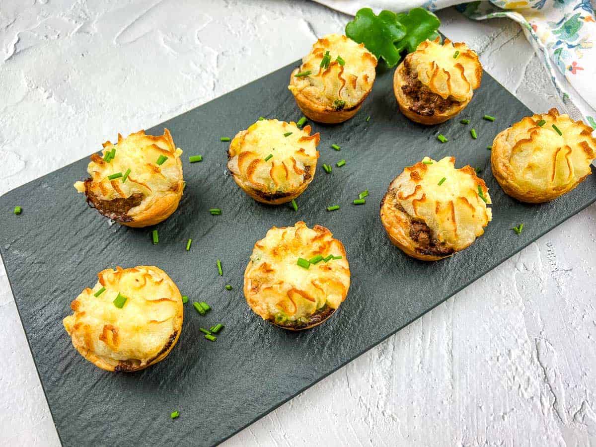 Eight Shepherd's Pie Bites are arranged on a slate board, garnished with chopped chives.