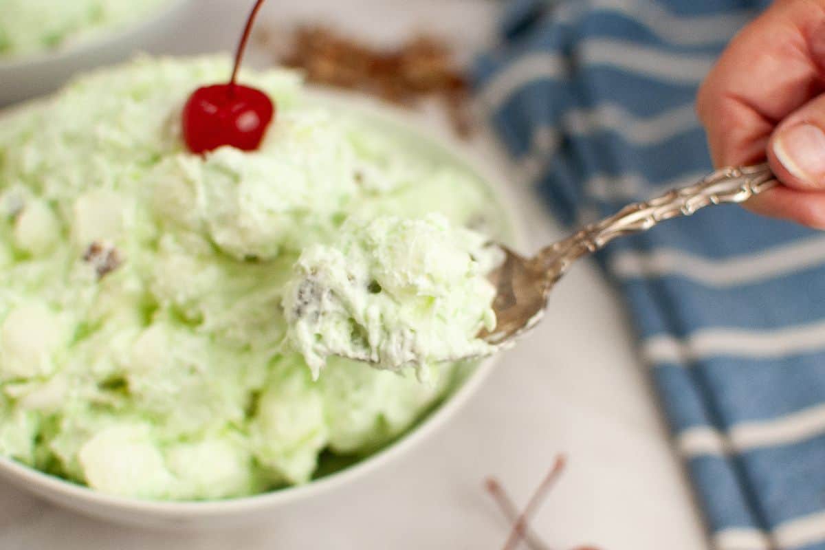 A spoonful of Watergate salad garnished with a cherry, is held over a bowl.