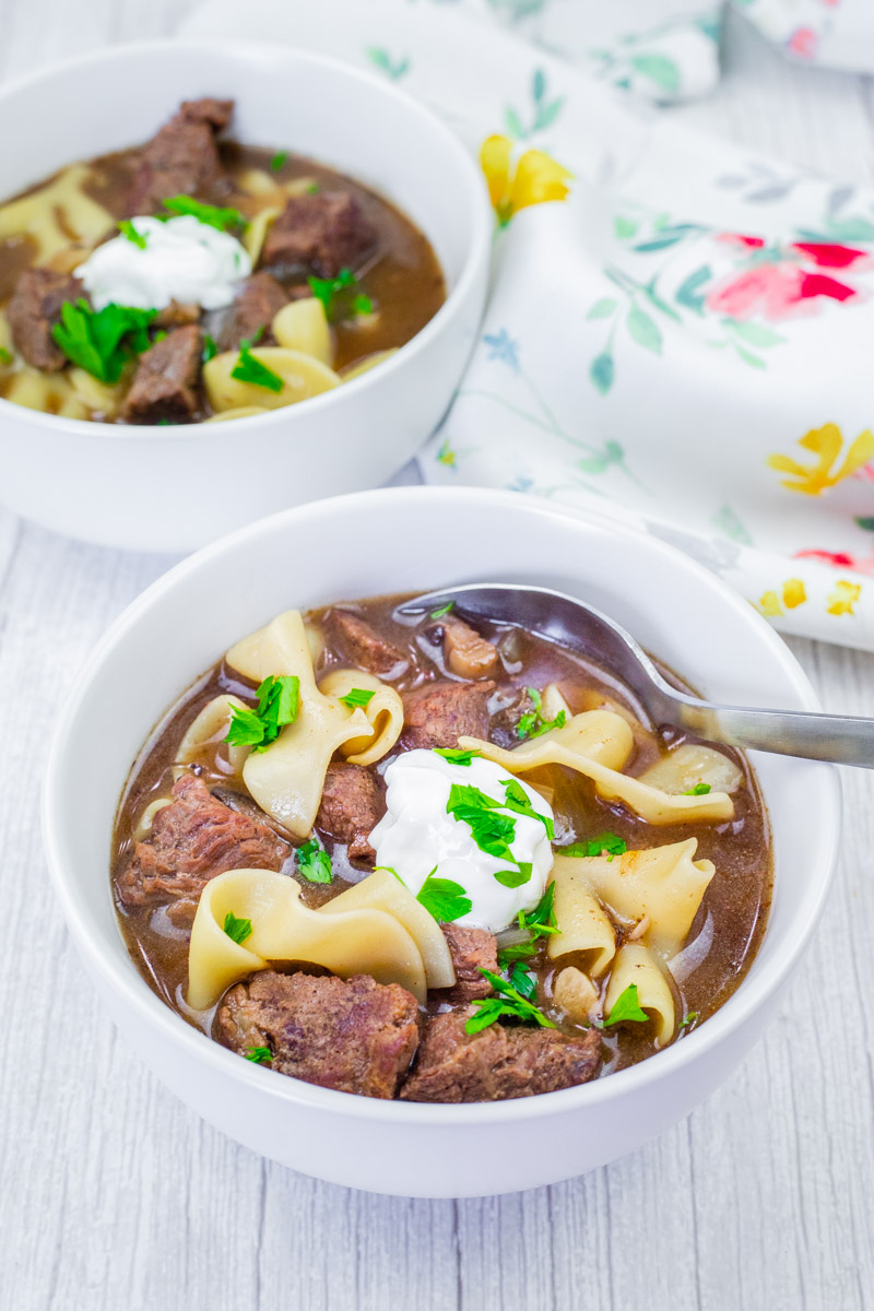 Two bowls of Beef Stroganoff Soup, garnished with sour cream and parsley, on a white table. A floral napkin is beside the bowls.