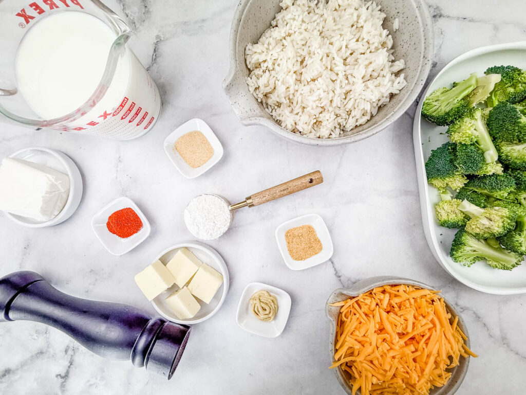 Ingredients for Broccoli Rice Casserole with No Canned Soup are arranged, including milk, cooked rice, broccoli, cheddar cheese, butter, cream cheese, red spice, flour, mustard, garlic powder, and a pepper grinder.