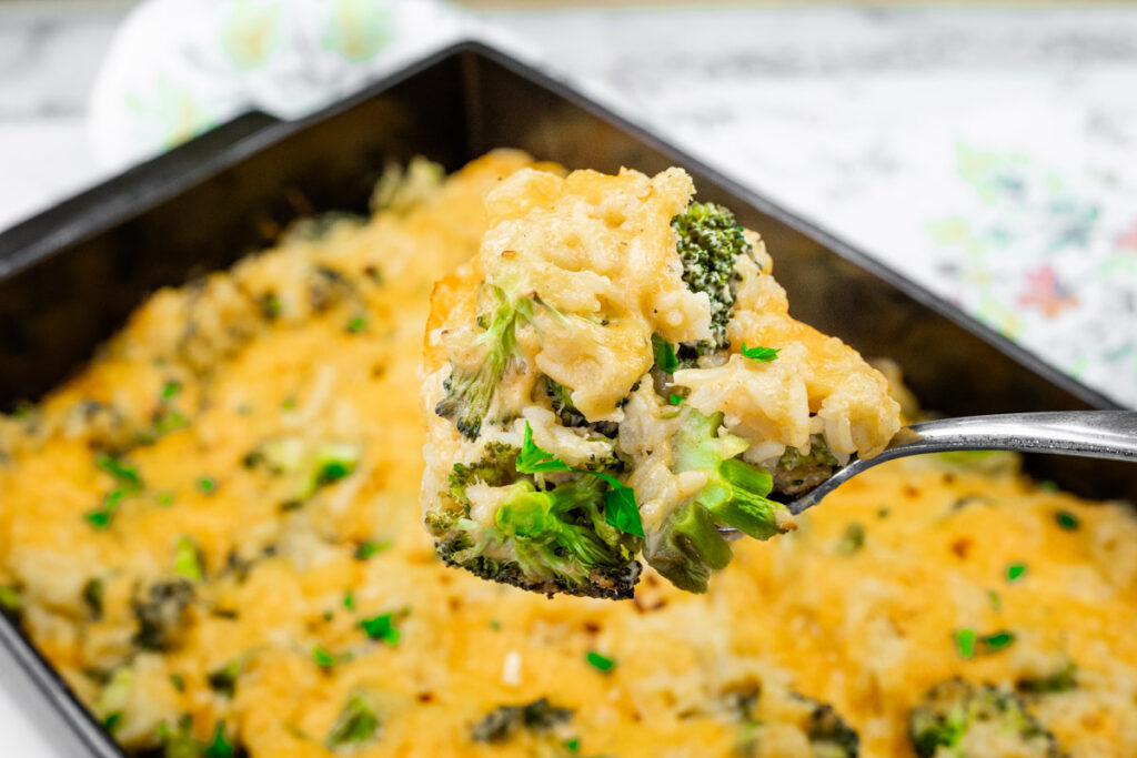 A close-up of a spoon holding a portion of Broccoli Rice Casserole with No Canned Soup, topped with melted cheese, over a baking dish filled with the same dish.