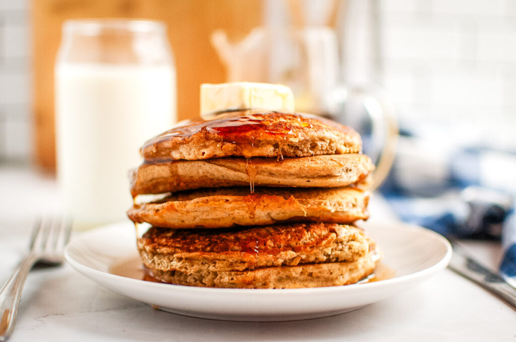 A plate of pancakes topped with butter and syrup, with a glass of milk in the background.