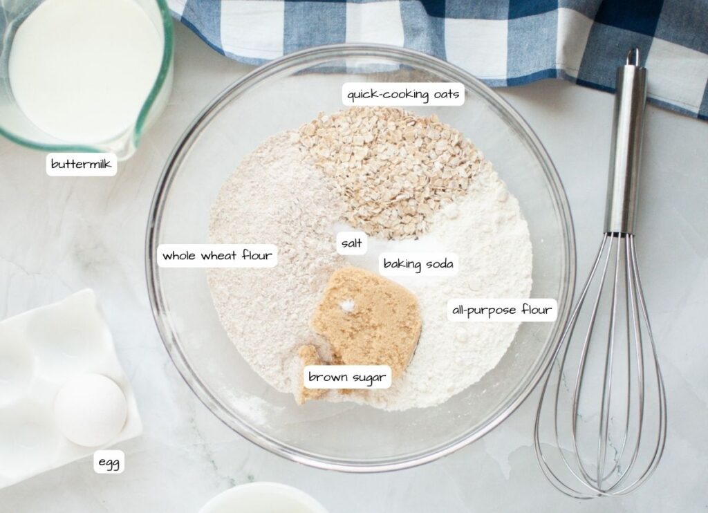 Labeled ingredients for buttermilk oatmeal pancakes, including oats, whole wheat flour, all-purpose flour, brown sugar, baking powder, and salt, surrounded by a whisk, a measuring cup of buttermilk, and an egg on a white marble surface with a blue-and-white checkered napkin.