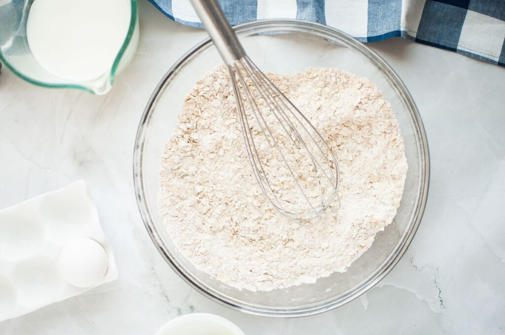 A glass bowl with whisked flour mixture on a marble surface, surrounded by a measuring cup of milk, an egg, and a blue checkered cloth.