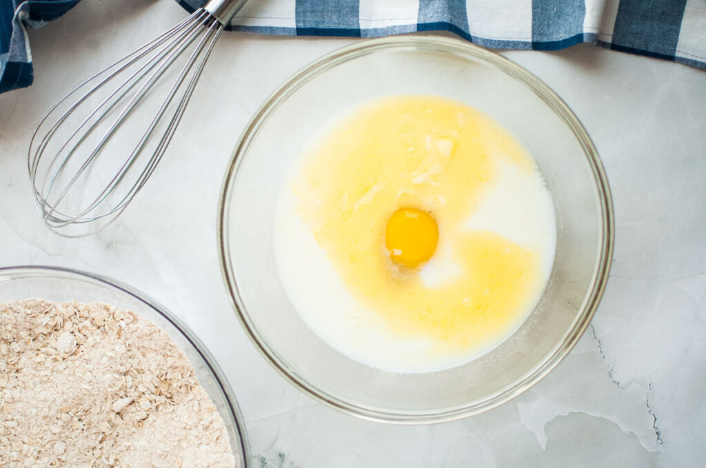 A bowl with an egg, milk, and butter mix, next to a whisk and a separate bowl of dry ingredients on a marble surface.