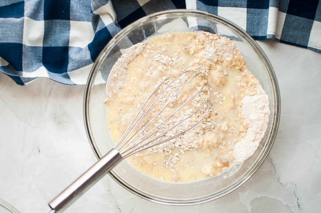 Mixing bowl with flour, oats, and liquid being whisked. Blue and white checkered cloth nearby on a marble surface.