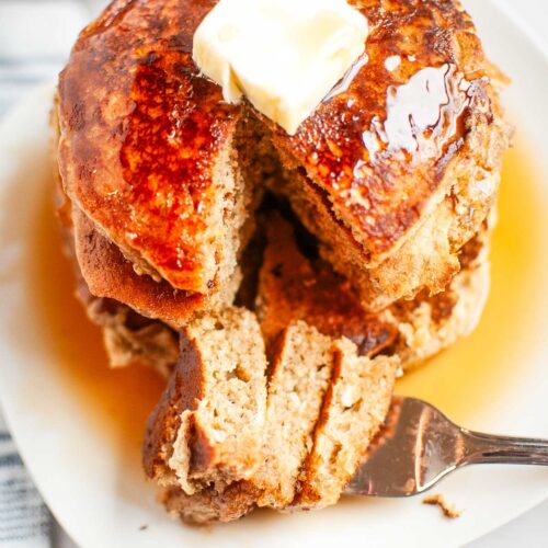 A stack of pancakes with butter on top and syrup drizzled, partially cut with a fork on a white plate.