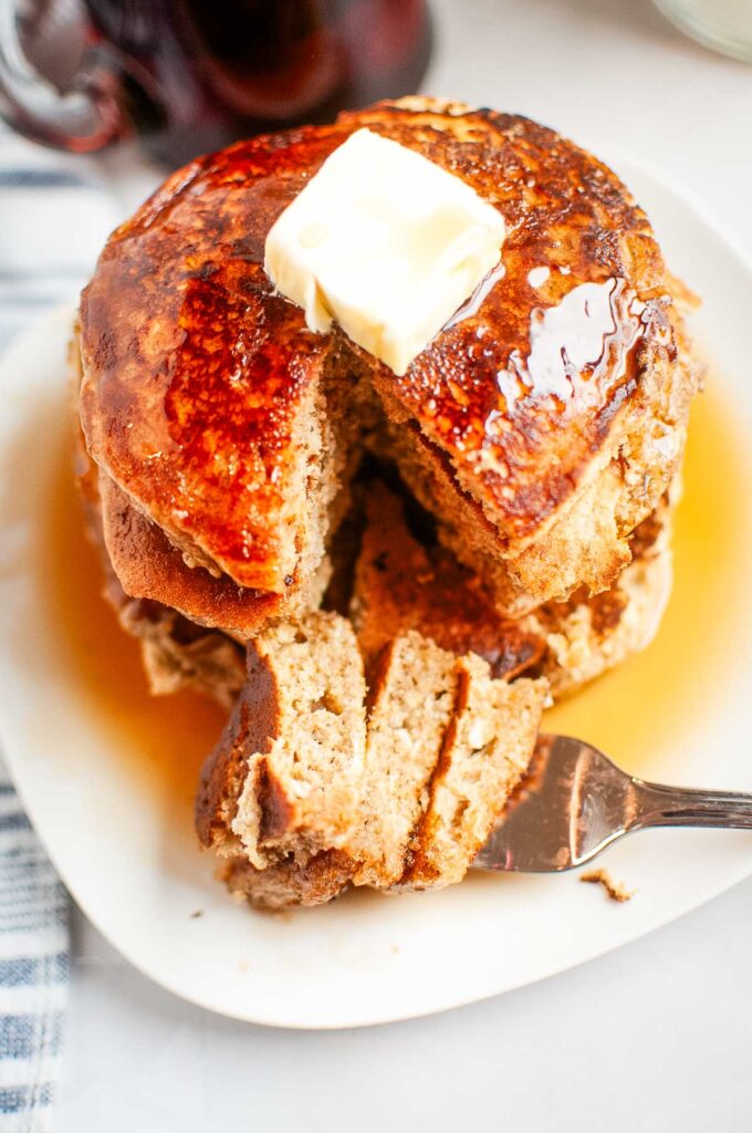 A stack of pancakes with butter on top and syrup drizzled, partially cut with a fork on a white plate.
