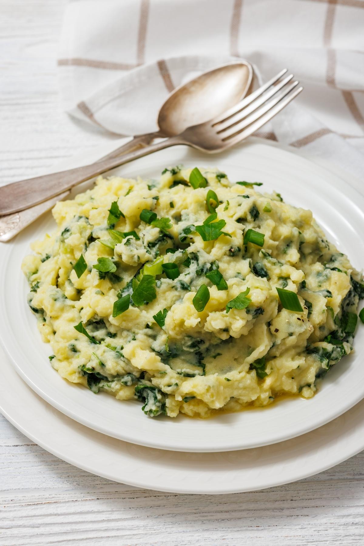 A plate of Colcannon with Kale, garnished with chopped green onions. A fork and spoon rest on a napkin beside the plate.