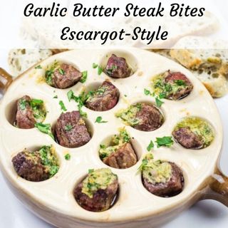 Ceramic dish with garlic butter steak bites garnished with herbs in escargot-style wells, accompanied by slices of bread.