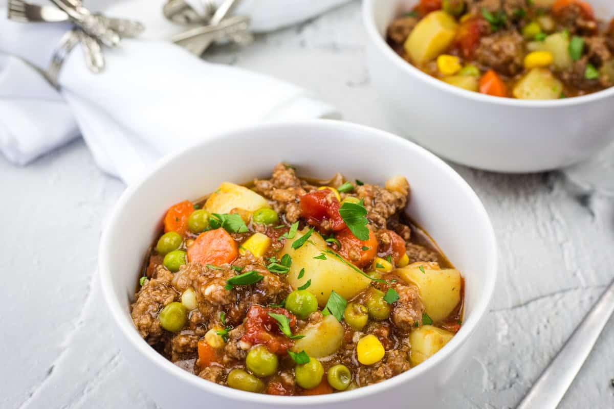 Two bowls of Homestyle hamburger stew with chunks of potato, carrots, peas, corn, and parsley garnish.