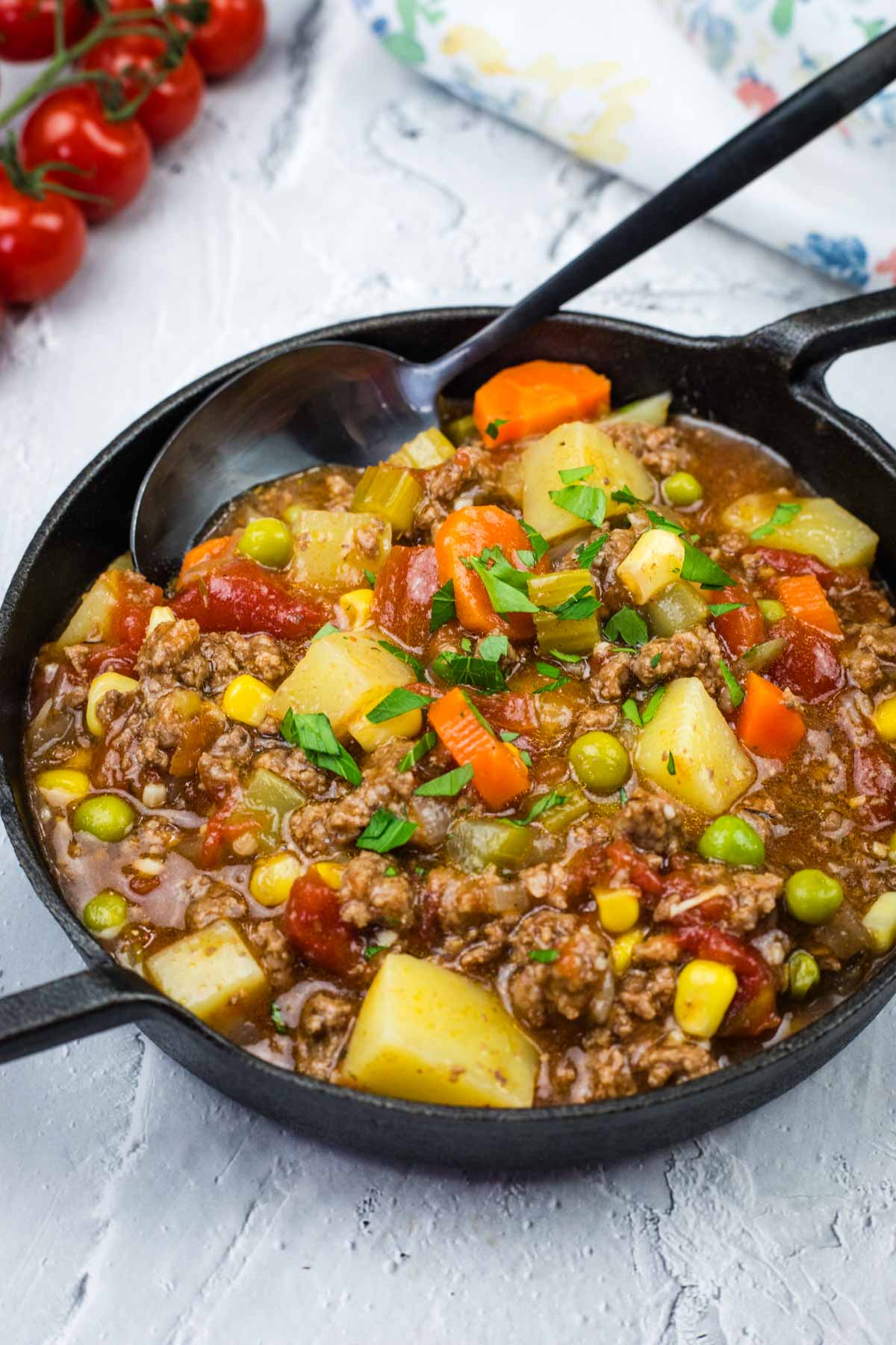 A skillet of classic hamburger stew containing ground meat, potatoes, peas, carrots, corn, and garnished with parsley. A spoon rests in the skillet.