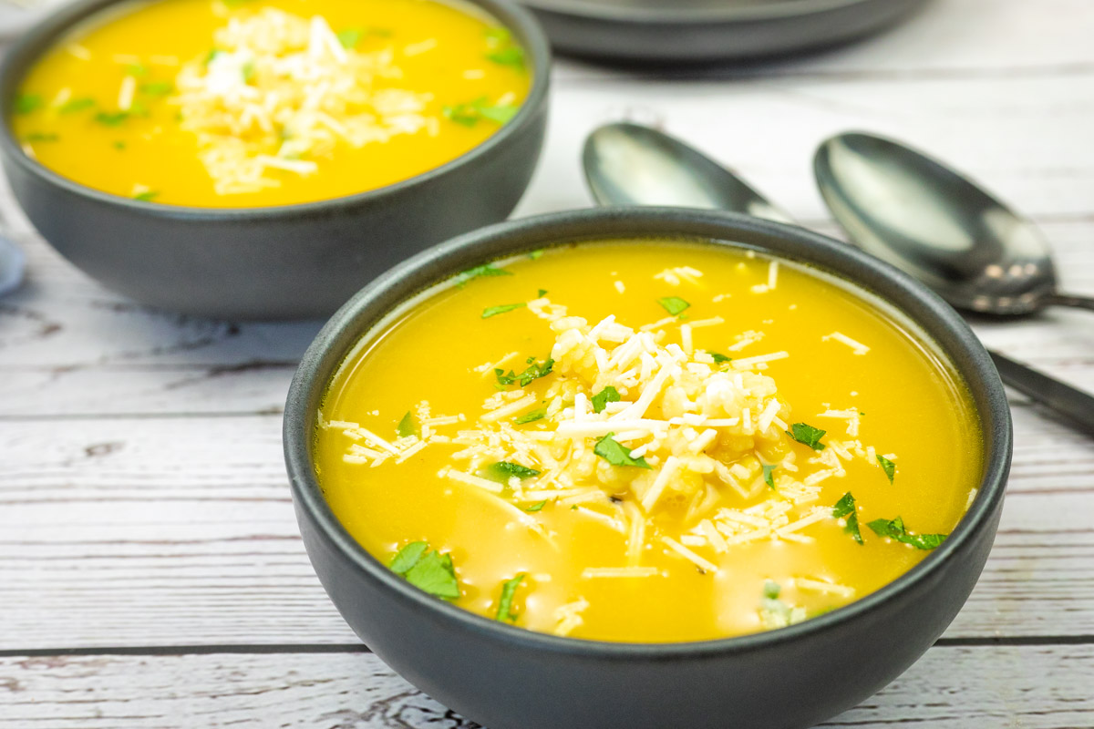 Bowls of Easy Pastina Soup garnished with shredded cheese and herbs on a wooden table, with spoons beside them.