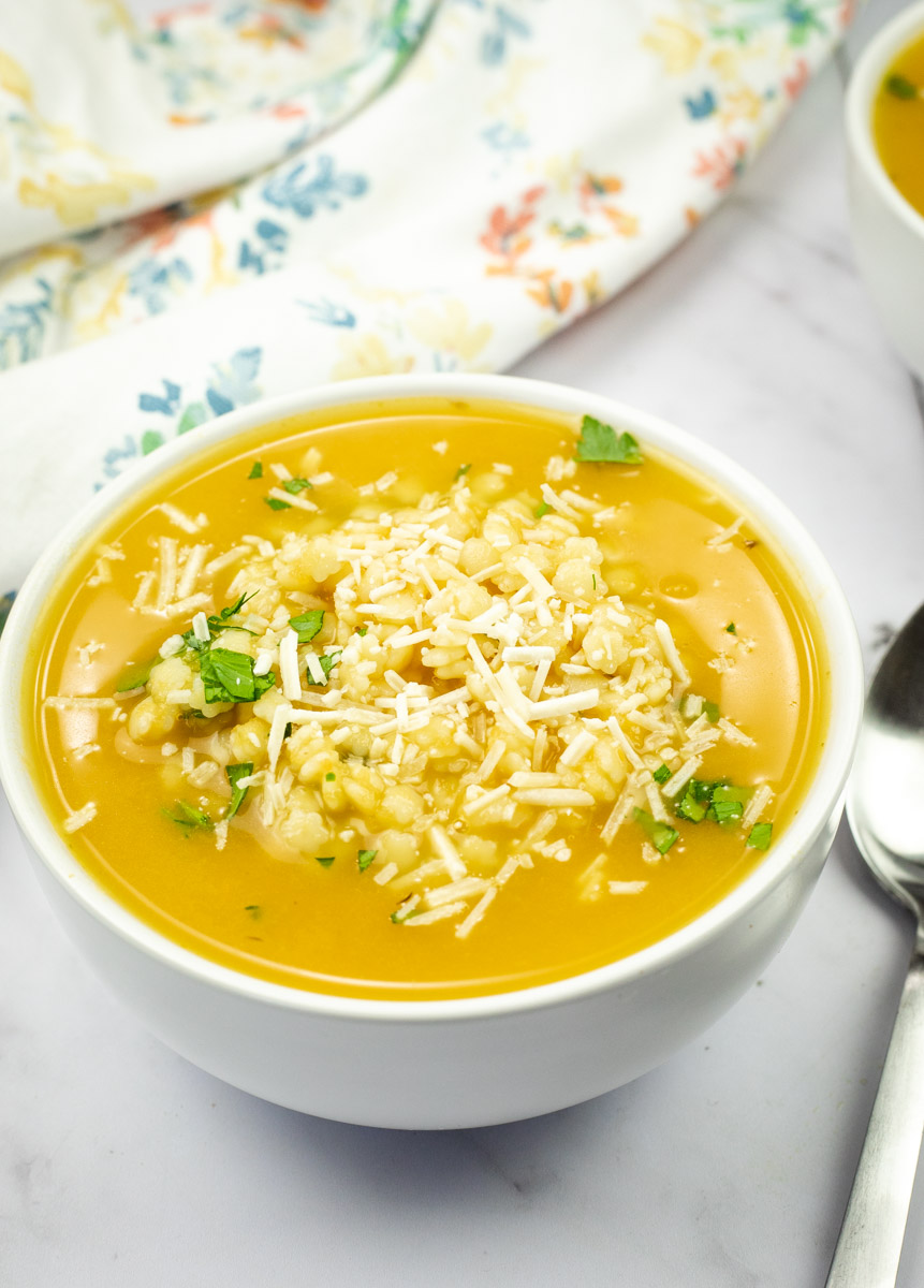 A bowl of pastina soup with shredded cheese and herbs on top, placed on a marble surface alongside a spoon.