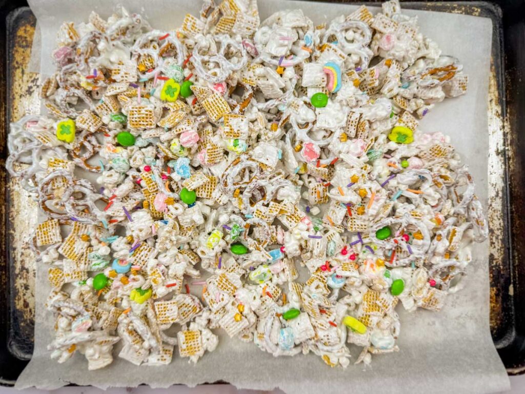 A tray of dessert mix containing pretzels, cereal squares, colorful candies, and white coating spread on parchment paper.