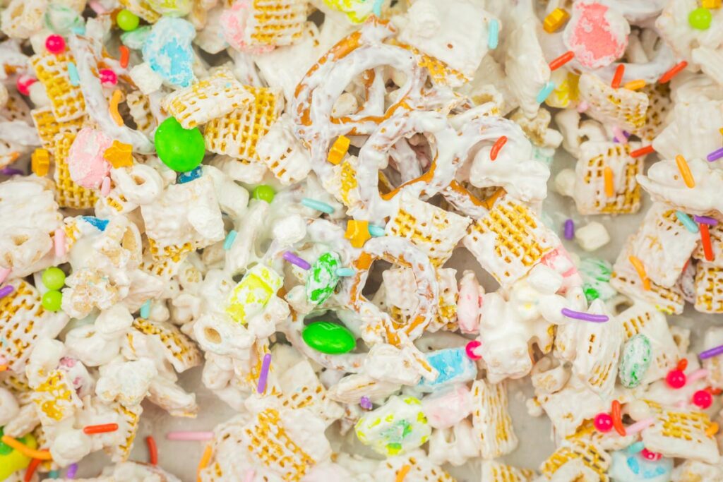 Colorful snack mix with pretzels, cereal, and candies coated in white chocolate, sprinkled with rainbow sprinkles.