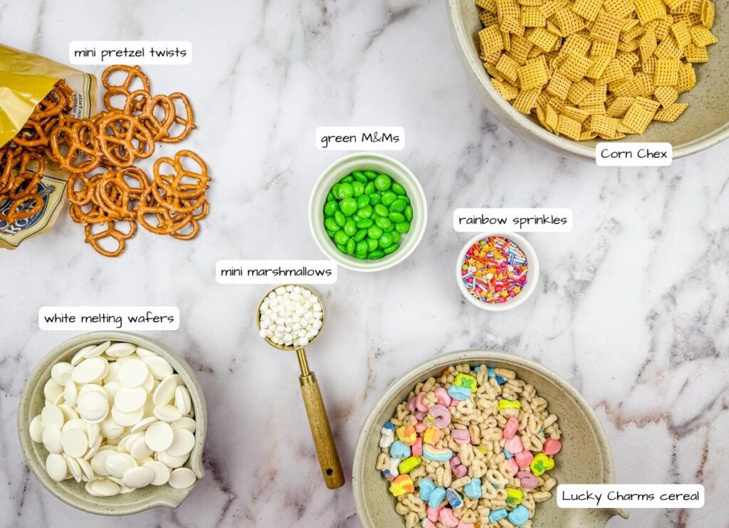 Ingredients for a snack mix include mini pretzel twists, corn chex, white melting wafers, green M&Ms, mini marshmallows, rainbow sprinkles, and Lucky Charms cereal on a marble surface.