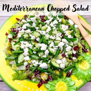 A Mediterranean chopped salad with cucumbers, lettuce, green beans, feta cheese, and radicchio on a yellow plate, garnished with wooden utensils.