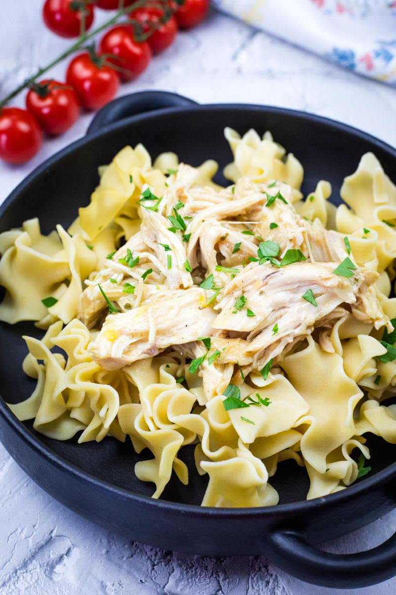Black bowl filled with egg noodles topped with shredded Mississippi Chicken - Slow Cooker, garnished with fresh parsley. A vine of cherry tomatoes rests in the background on a textured white surface.