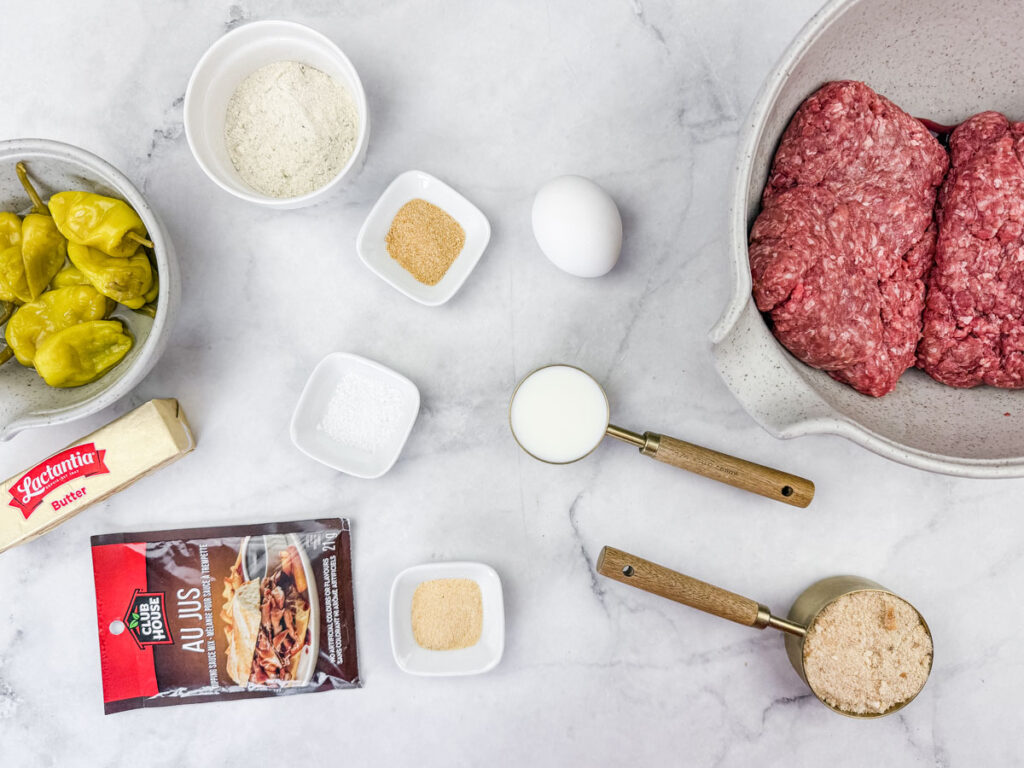 Ground beef in a bowl, surrounded by pepperoncini, flour, garlic powder, egg, sugar, milk, brown sugar, au jus packet, and butter on a marble surface.